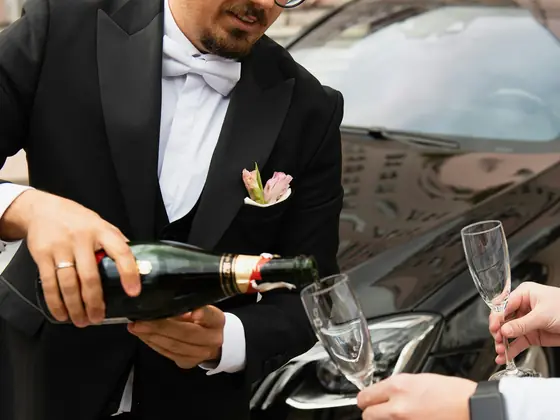 Man in a tuxedo pouring champagne beside a black luxury car